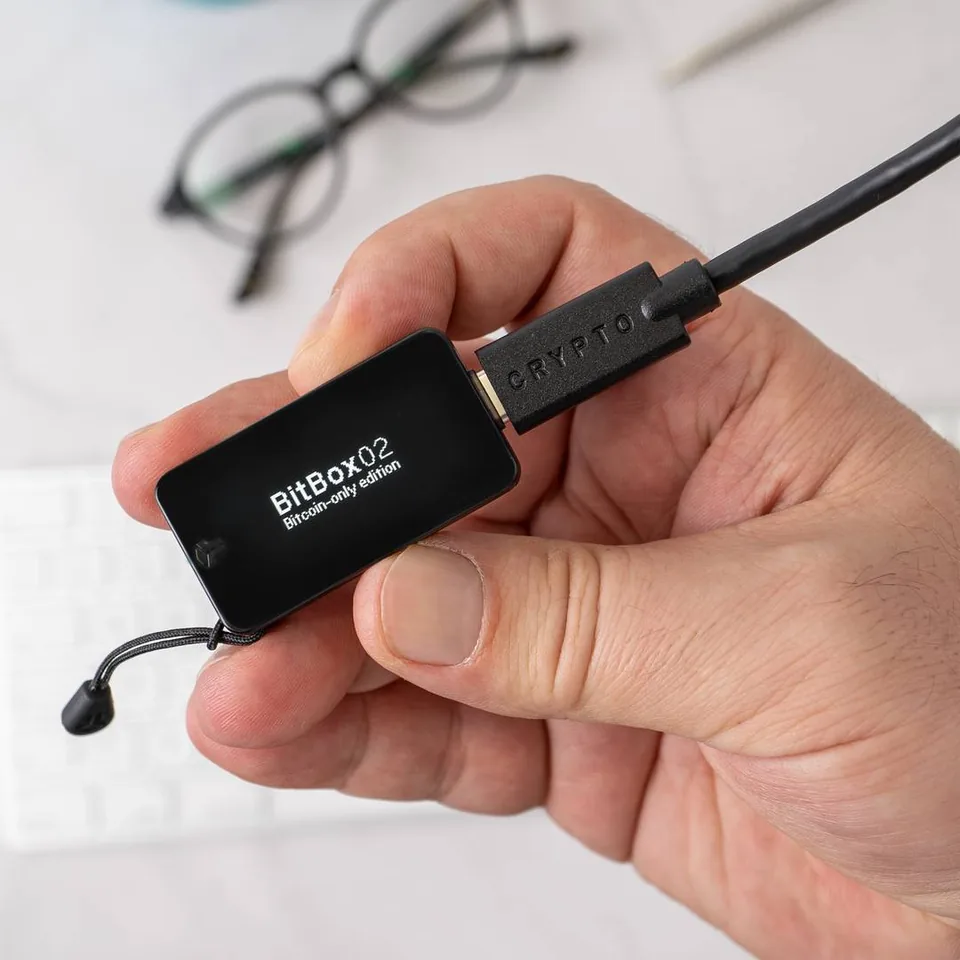 Manufacturer photograph of an original BitBox02 held at its edges, connected to a USB cable. Its screen identifies the Bitcoin-only edition.