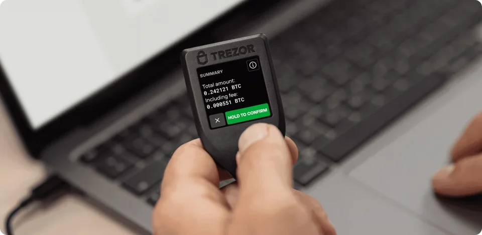 A Trezor Model T held beside a laptop, with transaction details on the device screen.