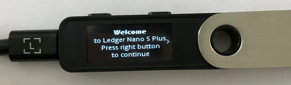 Ledger photograph of Nano S Plus connected by USB, with its welcome screen and two buttons above the display visible.