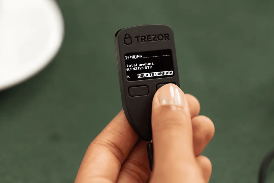 A hand holding a connected Trezor Model One with a transaction confirmation prompt on its screen.