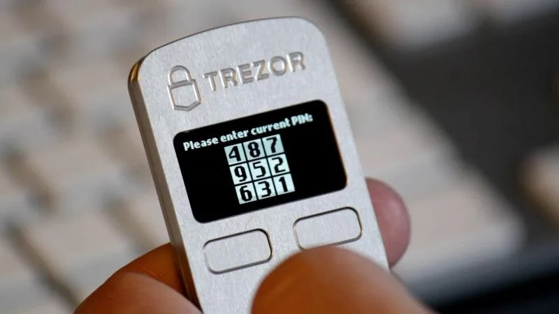 A Trezor Model One held in a hand, displaying a shuffled nine-position PIN grid.