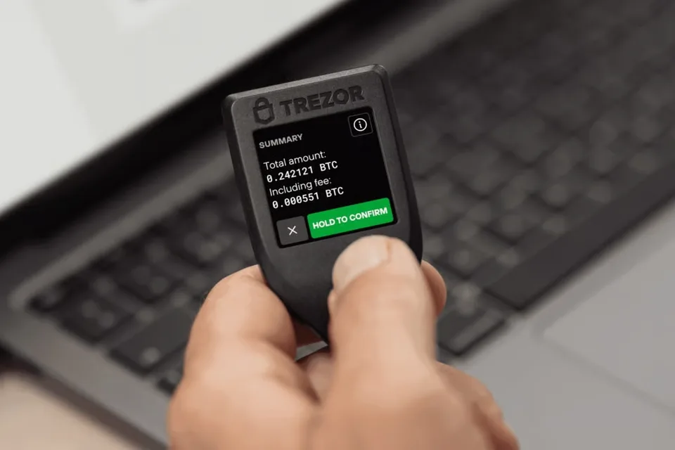 A hand holds Trezor Model T beside a laptop. Its touchscreen shows a Bitcoin transaction summary and Hold to confirm.