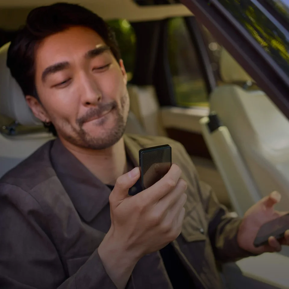 Manufacturer photograph of a seated person holding a dark Trezor Safe 7 with its rear facing the camera and a smartphone separately in the other hand.