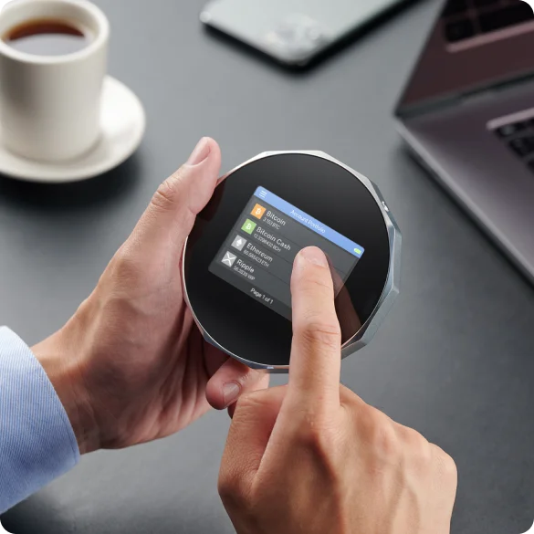 Hands holding a round SecuX V20 beside a laptop, with a finger touching the account list on its screen.