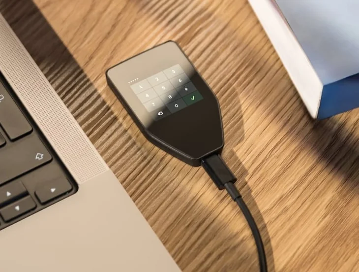 Trezor Safe 5 beside a laptop, with a USB cable attached and a PIN keypad visible on its touchscreen.
